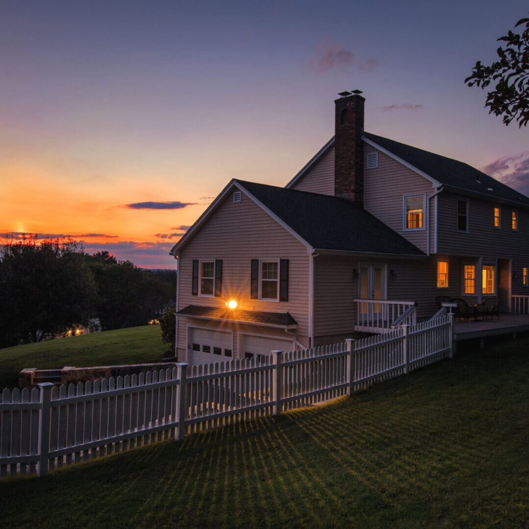 A peaceful house at sunset with glowing windows and a white fence.